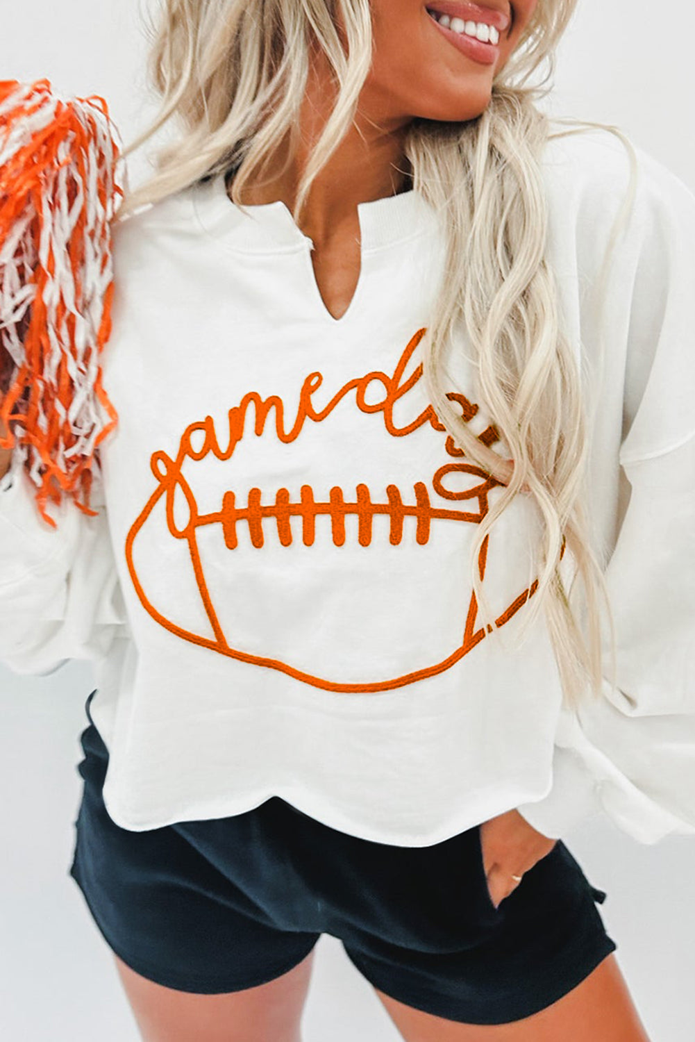 A woman with long blonde hair dressed in a chic White Game Day Lettering Rugby Football Notched Neck Sweatshirt and blue jeans stands near a Christmas tree adorned with red and white ornaments and ribbon.