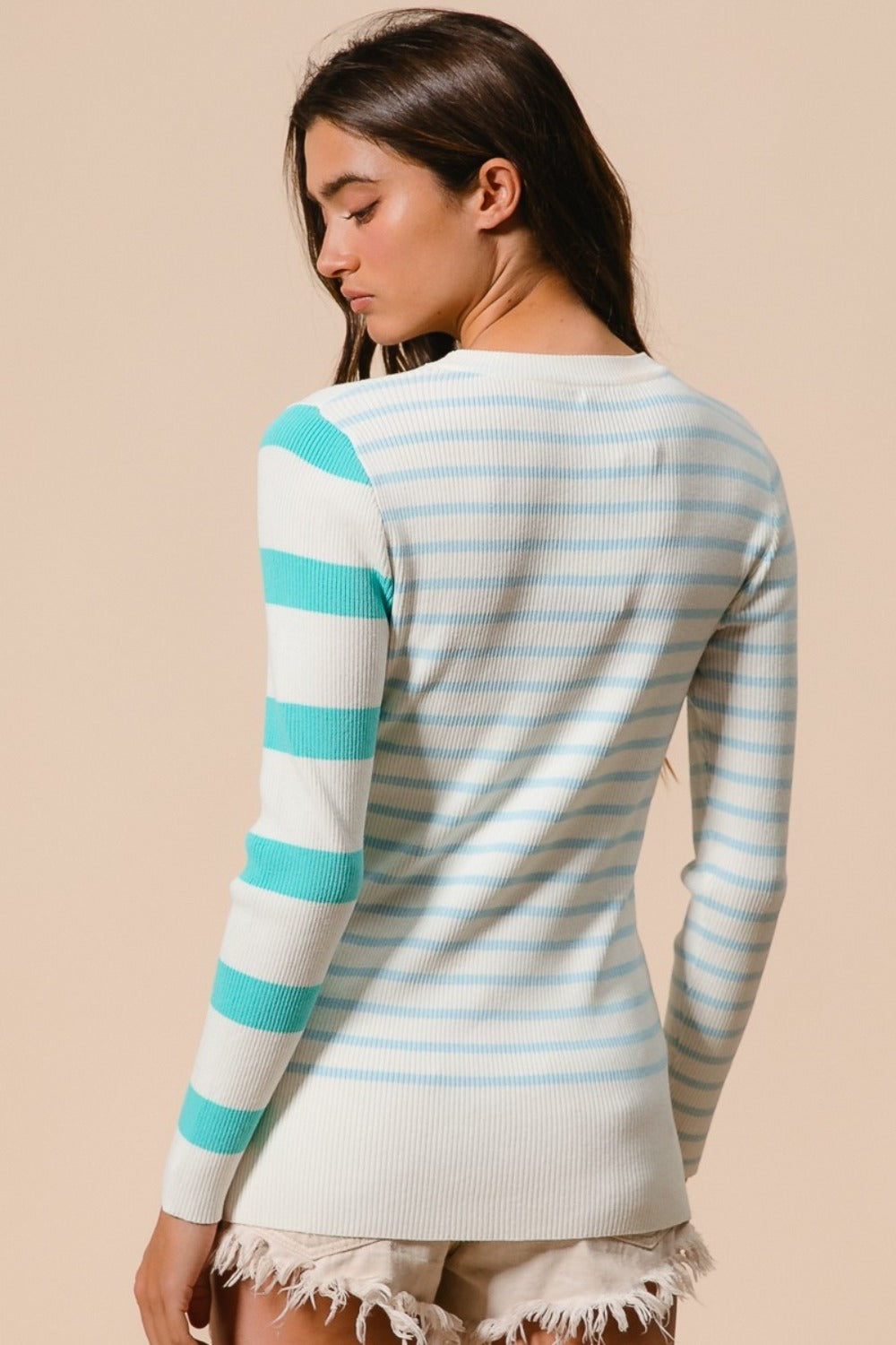 A person with long hair is wearing a BiBi Contrast Striped Asymmetrical Hem Knit Top featuring bold white and teal stripes, paired with white distressed shorts, and stands against a beige background.