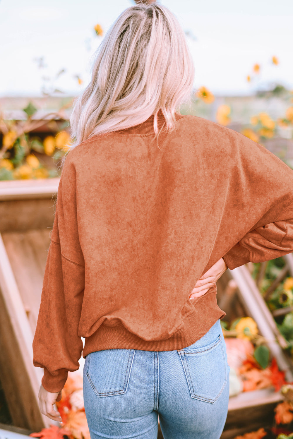 A person with long blonde hair wearing an Orange Drop Shoulder Crew Neck Pullover Sweatshirt and blue jeans stands with their back to the camera in a sunflower field.