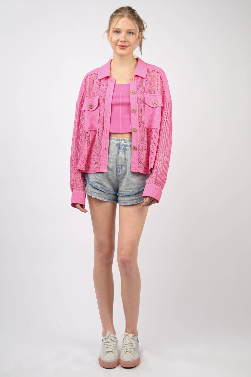 A person wearing a bright pink, long sleeve button-up lace shirt with drop shoulders and high-waisted denim shorts stands against a plain background.