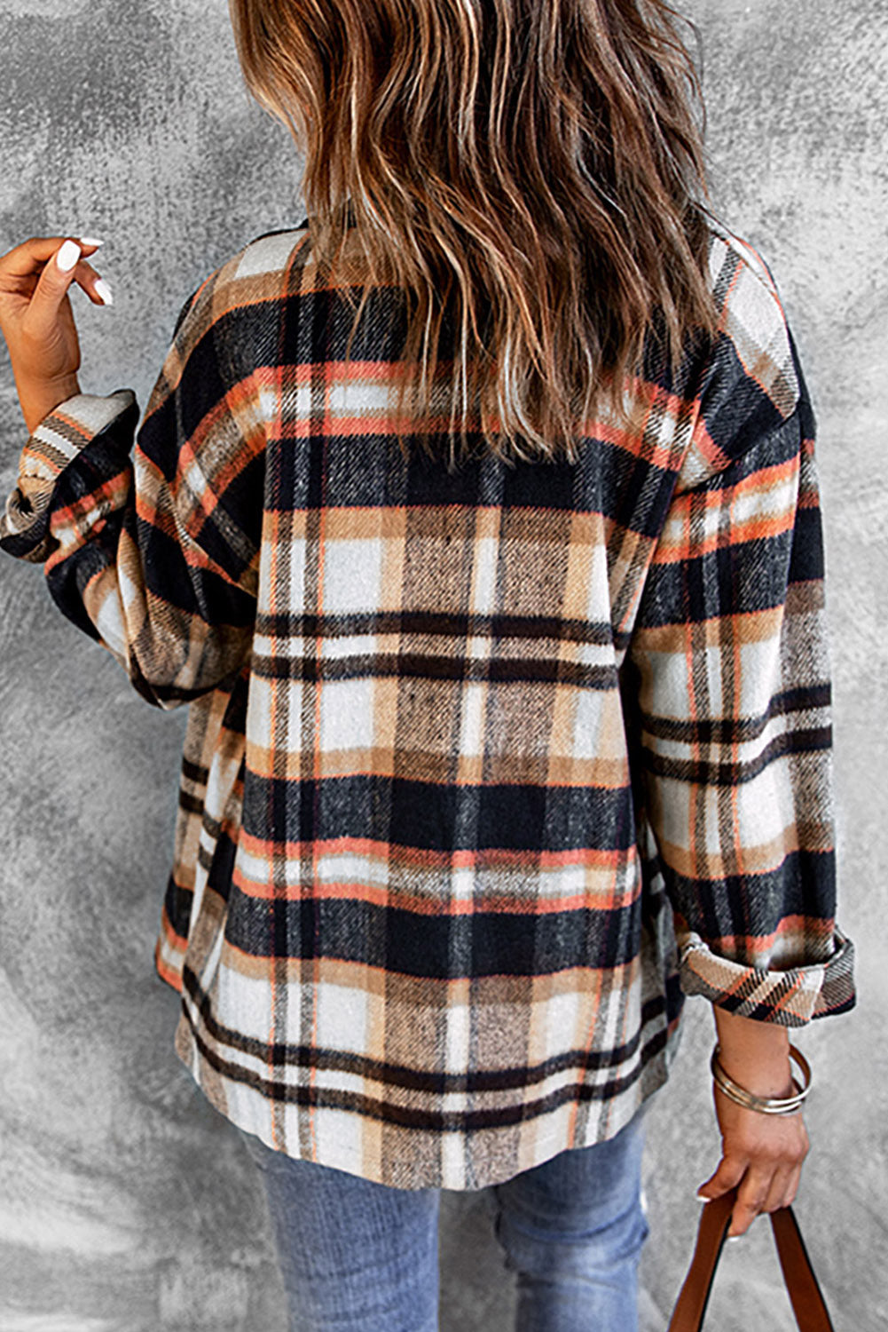 A person wearing a Brown Plaid Button Up Long Sleeve Flannel Shacket layered over a white top with ripped jeans and holding a brown handbag stands against a textured gray wall.