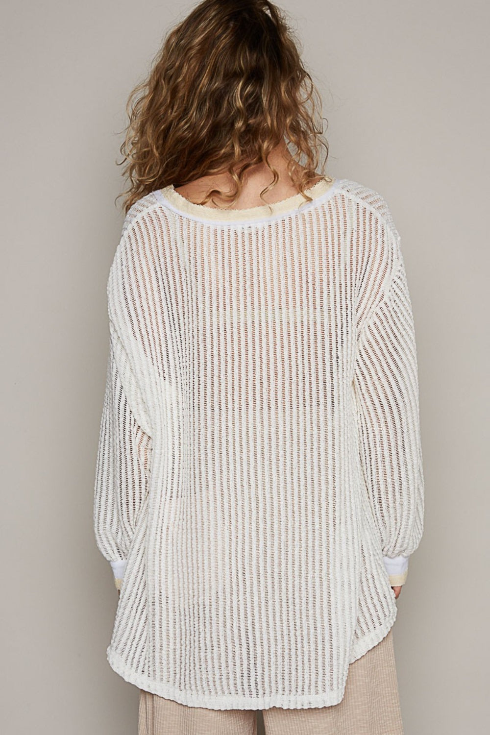 A person with wavy hair wearing a loose, bohemian-style POL V-Neck Long Sleeve Crochet Top in a sheer, striped white design paired with beige pants stands against a plain background.