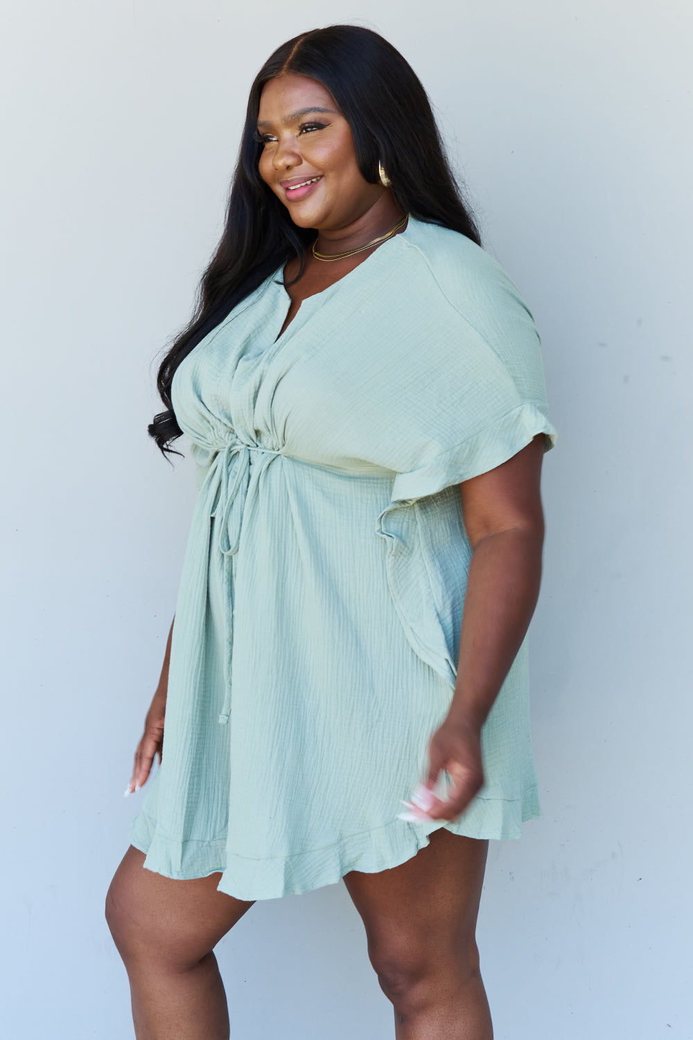 A woman is wearing the Ninexis Out Of Time Full Size Ruffle Hem Dress with Drawstring Waistband in Light Sage. Her long, dark hair falls gracefully as she smiles and looks to her left, standing against a plain background.