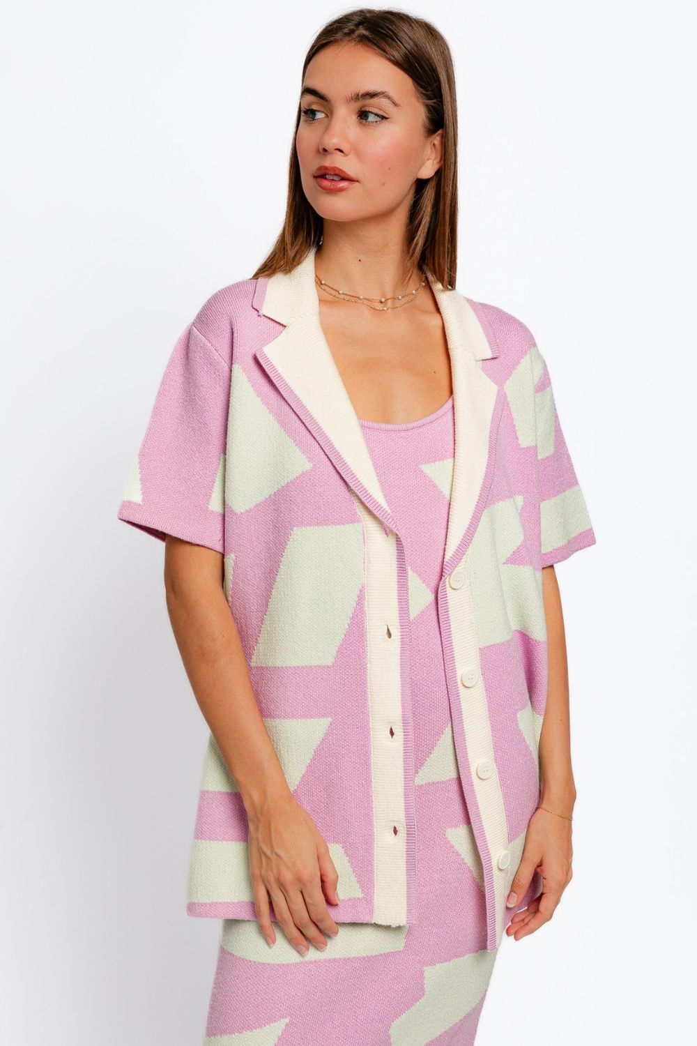 A woman is wearing the Le Lis Abstract Contrast Short Sleeve Collared Cardigan in pastel pink and cream with a geometric pattern, paired with a matching dress. She embodies a modern artistic touch as she looks to the side with her hands at her sides.