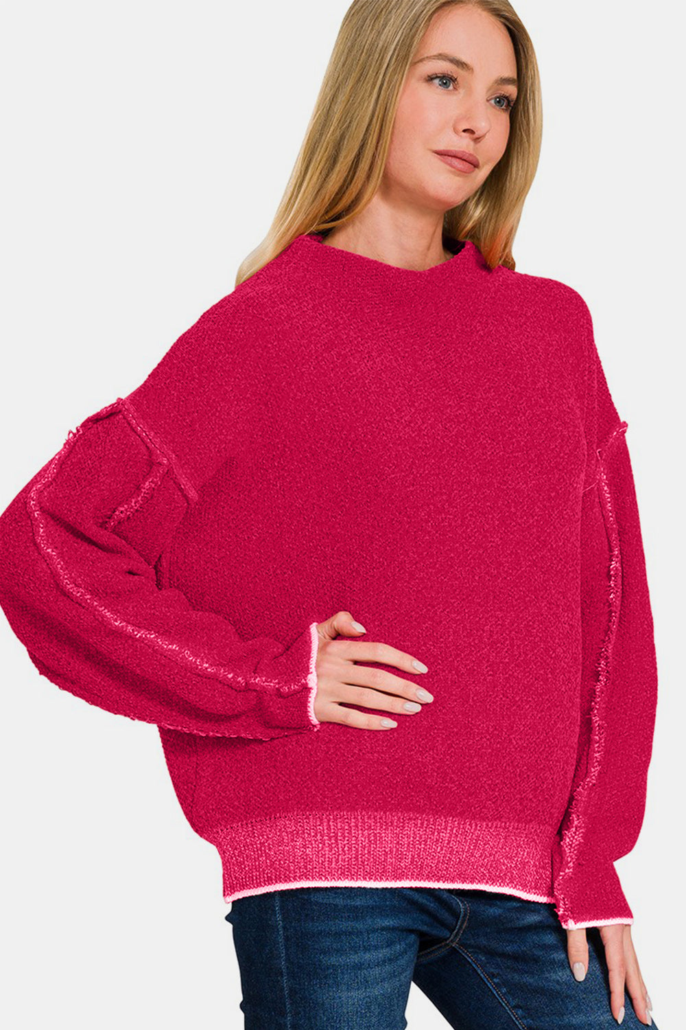 A woman in a vibrant pink Zenana Exposed Seam Mock Neck Long Sleeve Sweater and jeans stands elegantly against a minimalist white background.