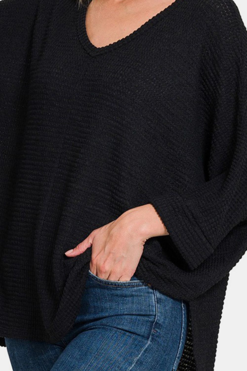 A woman wearing the Zenana Full Size Waffle Knit V-Neck Long Sleeve Slit Top paired with blue jeans stands with one hand touching her head and the other in her pocket.