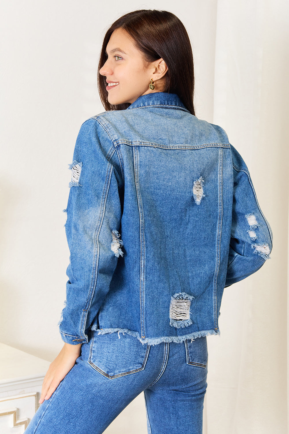 A person smiles while wearing the Baeful Leopard Floral Distressed Raw Hem Denim Jacket with embroidered patches, paired with a white shirt and blue jeans, as they stand indoors near a white piece of furniture.