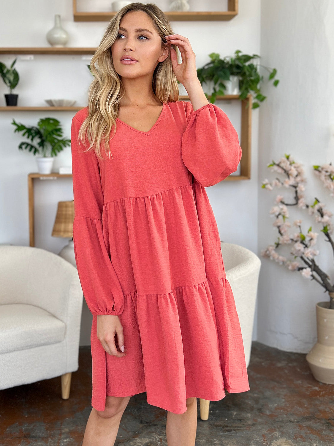 A woman stands wearing the Double Take Full Size V-Neck Balloon Sleeve Tiered Dress with Pockets in pink. Crafted from high stretch yarn, this dress combines comfort and style effortlessly. She has long, wavy hair and accessorizes with hoop earrings and a necklace. This imported garment is easy to care for – simply machine wash cold to keep it looking fresh.