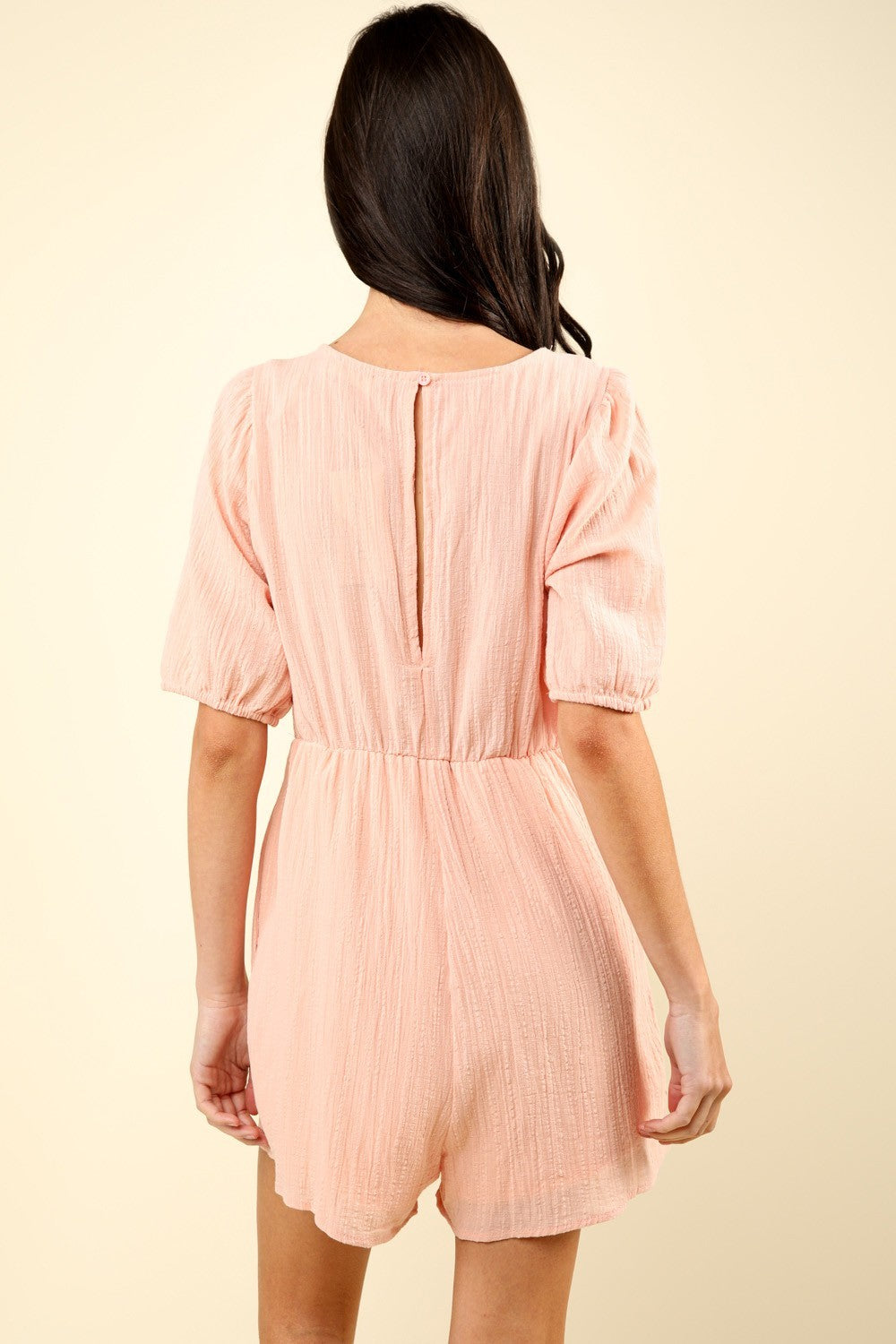 A woman in the VERY J Lace Detail Puff Sleeve Romper with Pockets, featuring light pink fabric and intricate lace detailing around the chest, stands against a plain beige background.