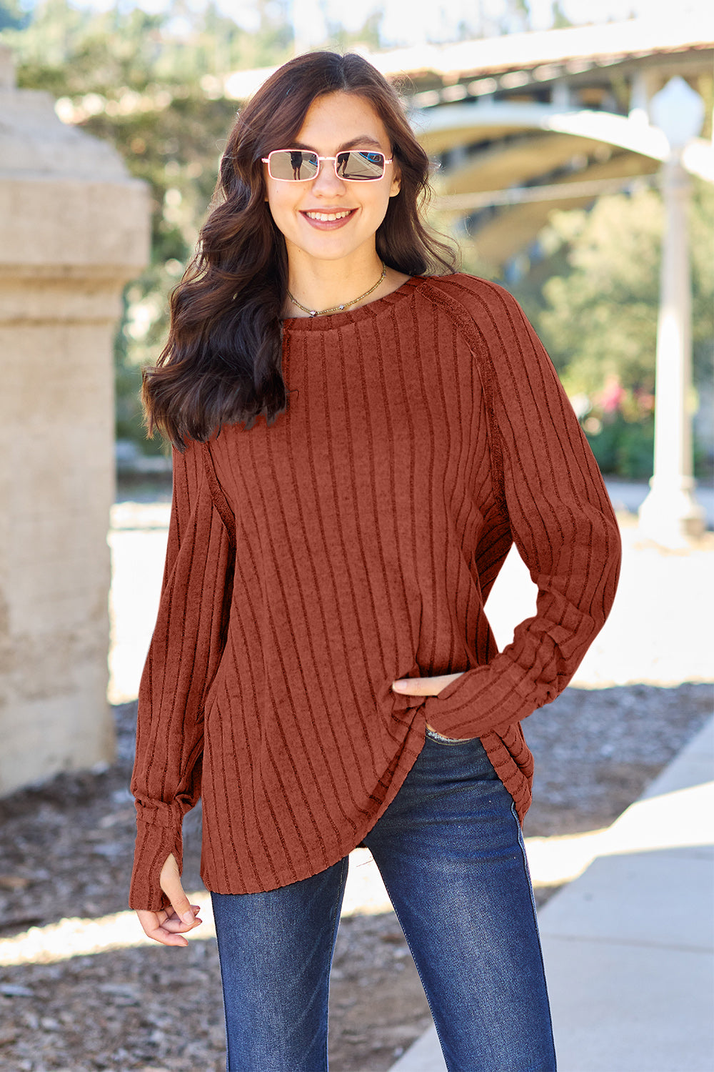 A person with long dark hair and sunglasses stands outdoors, wearing the Basic Bae Full Size Ribbed Round Neck Long Sleeve Knit Top in blue and a pair of jeans. The basic style outfit blends seamlessly with the background of trees and a bridge.