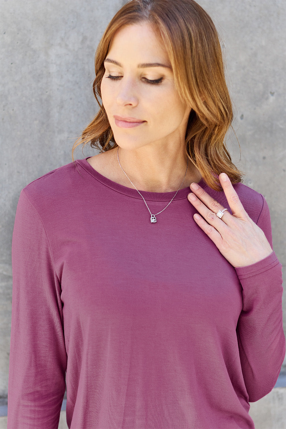 A woman with shoulder-length hair wearing the Basic Bae Full Size Round Neck Long Sleeve Top and blue jeans stands in front of a concrete wall, holding a beige handbag over her shoulder. Her outfit exemplifies basic style, with the top made from 100% polyester and recommended for machine wash cold.