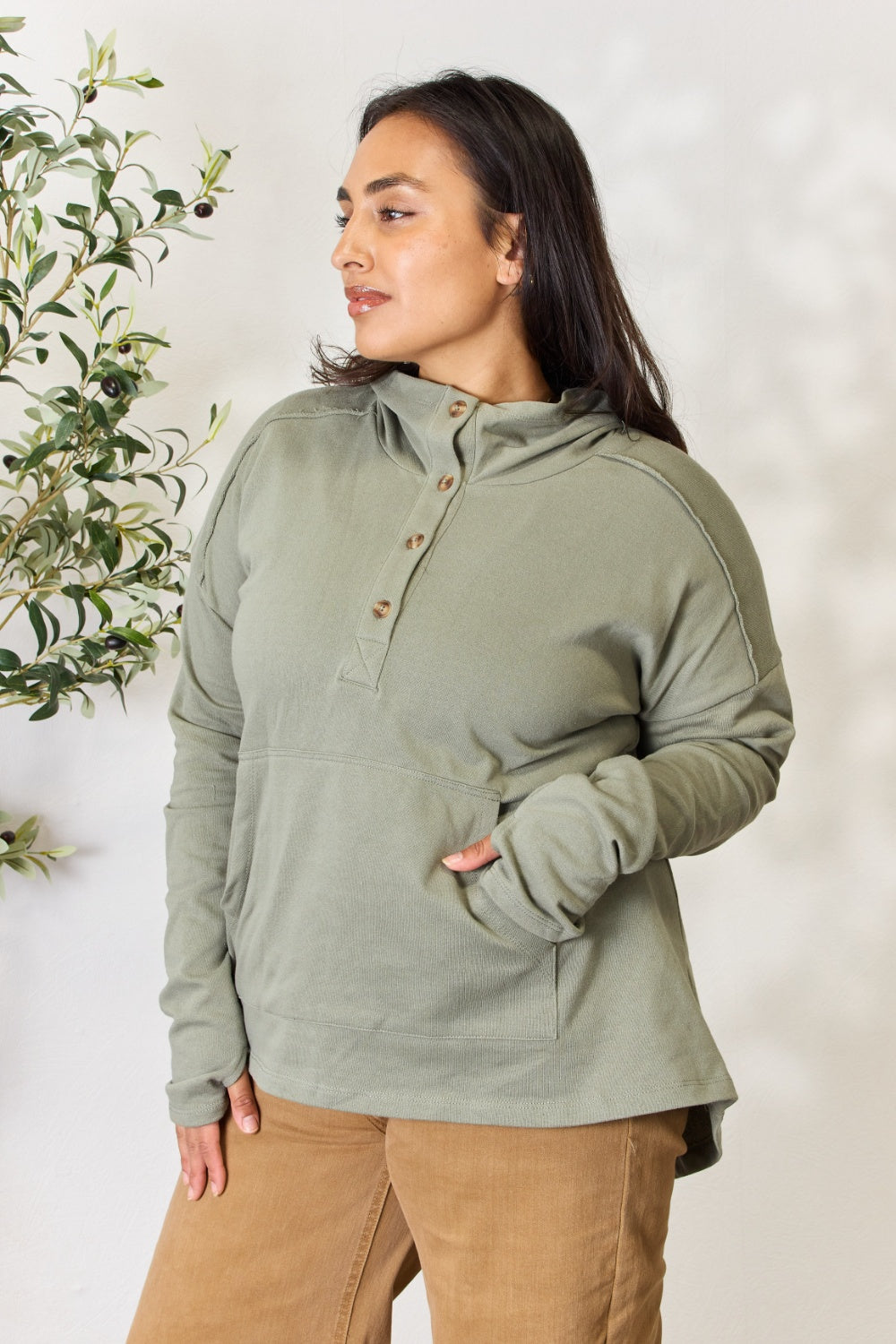 A person stands with hands in the pockets of their light green Culture Code Full Size Half Button Hoodie and brown pants. A plant is visible on the left side of the image.