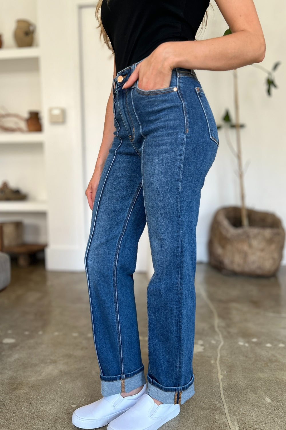 Two women stand side by side indoors, both wearing black tank tops, Judy Blue Full Size High Waist Front Seam Detail Straight Jeans, and white shoes. They are smiling and posing for the camera. A plant and shelves with decorations are in the background.