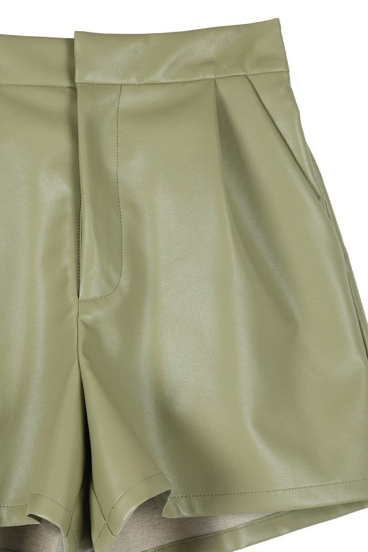 An individual is dressed in a light green sweater paired with pleated vegan leather shorts, featuring a functional pocket where one hand rests.