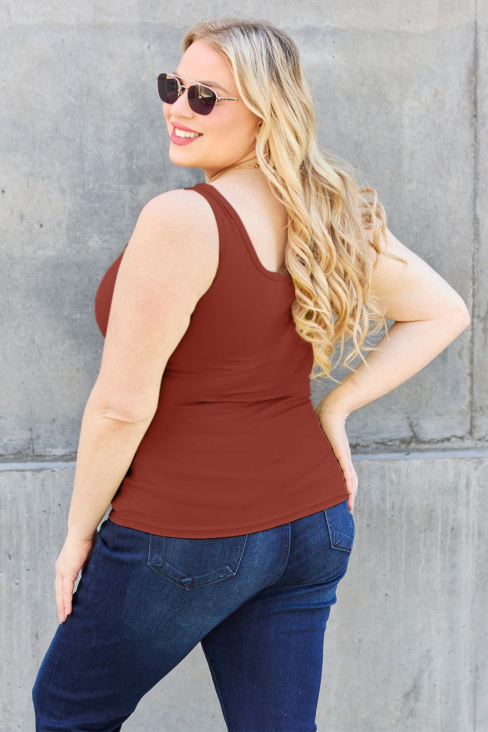 A woman with brown hair, wearing sunglasses, blue jeans, and the Basic Bae Full Size Square Neck Wide Strap Tank in black stands against a concrete wall, showcasing a basic style.