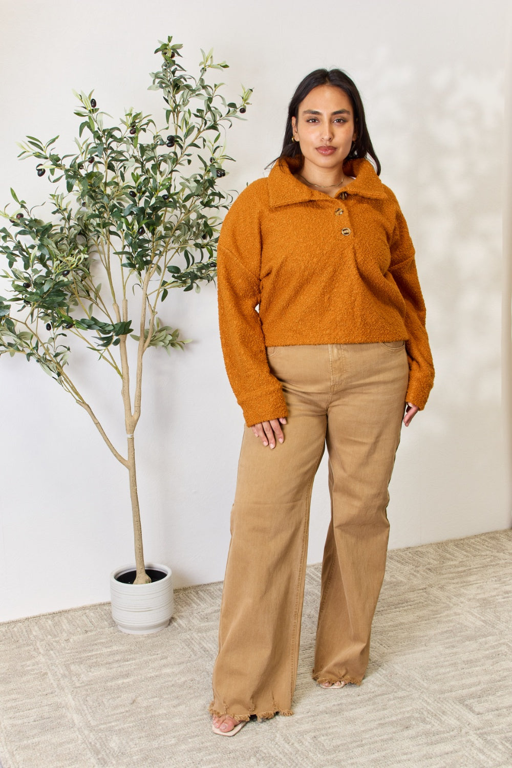 A person wears a brown textured Culture Code Full Size Half Button Turtleneck Sweatshirt made from breathable fabrics and tan pants, standing next to a leafy plant against a light background.