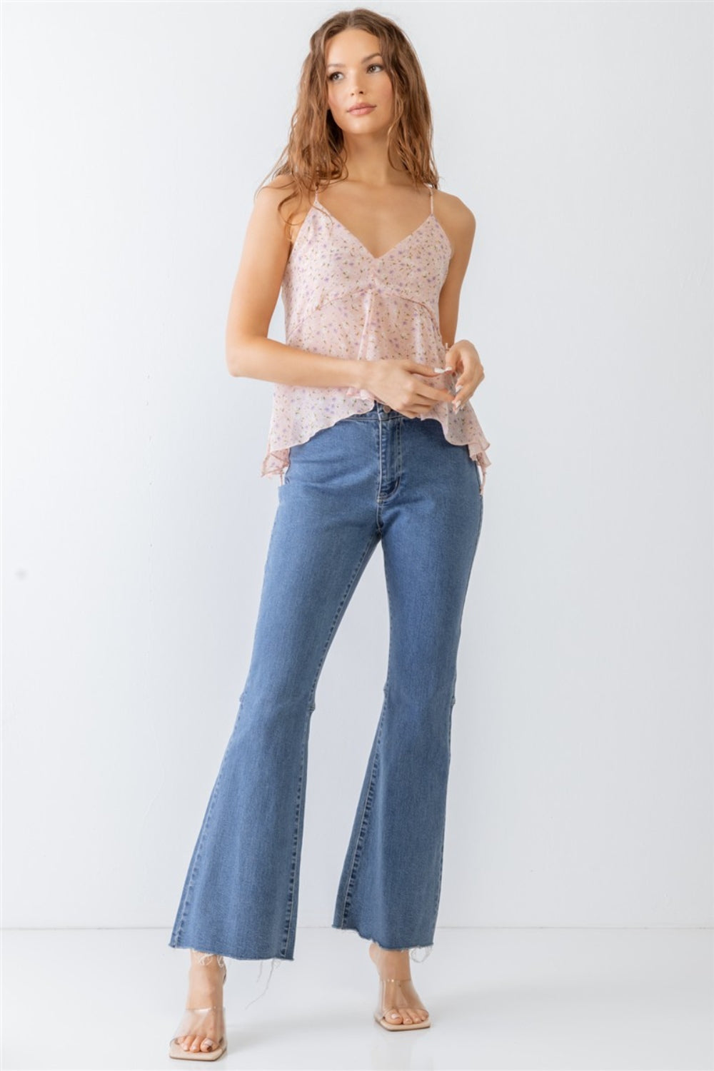 A woman wearing the Miss Love / Kevin Co. Floral Print V-Neck Criss-Cross Back Flare Cami and blue jeans stands against a plain white background.