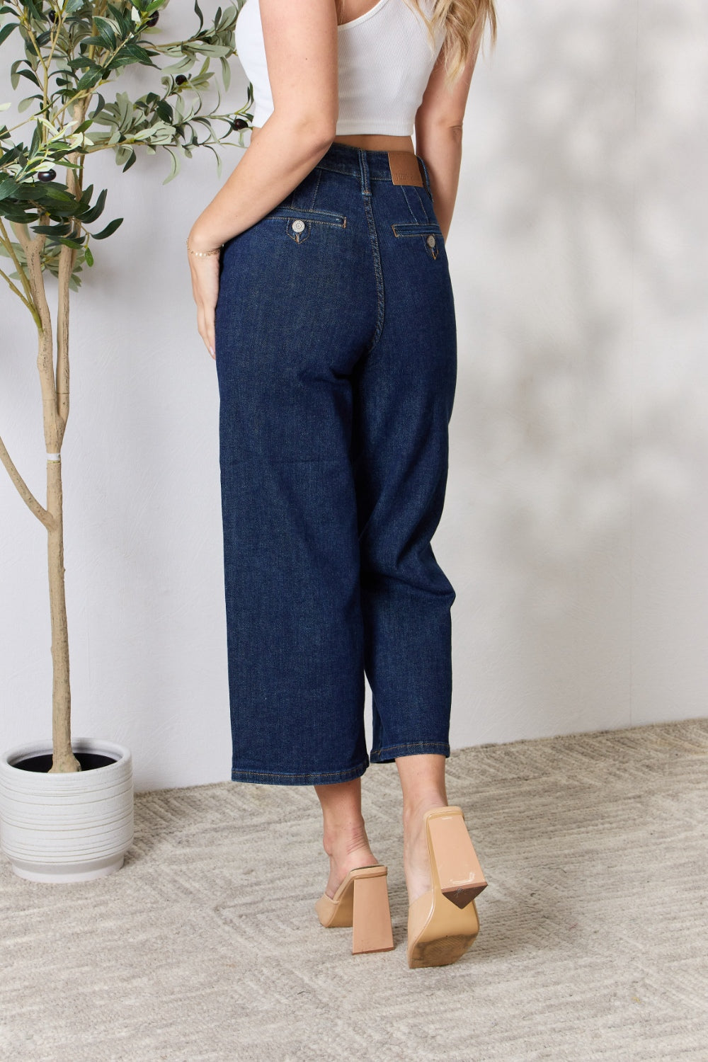 A person wearing the Judy Blue Full Size High Waist Cropped Wide Leg Jeans, which are renowned for their tummy control design, and nude block heel sandals stands near a potted plant.