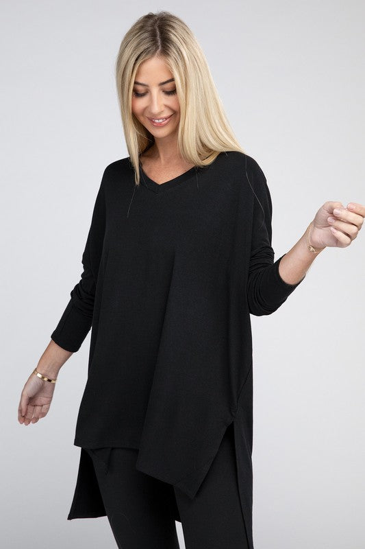 A woman with long, blonde hair is wearing a loose-fitting, brown Dolman Long Sleeve V-Neck Side Slit Hi-Low Hem Top and black pants. She poses with one hand on her hip and a neutral facial expression against a light grey background.