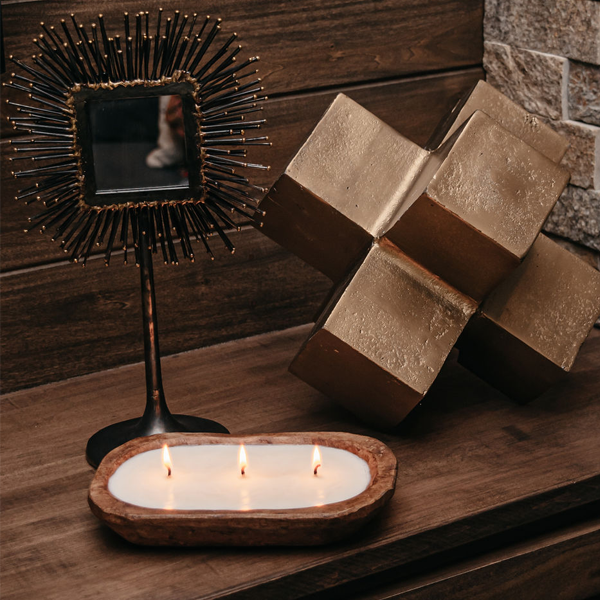 A three-wick dough bowl soy candle named "Pillow Talk" rests on a wooden surface, surrounded by lush green foliage and delicate rose petals. The scent features notes of peony, jasmine, rose, and lush suede.