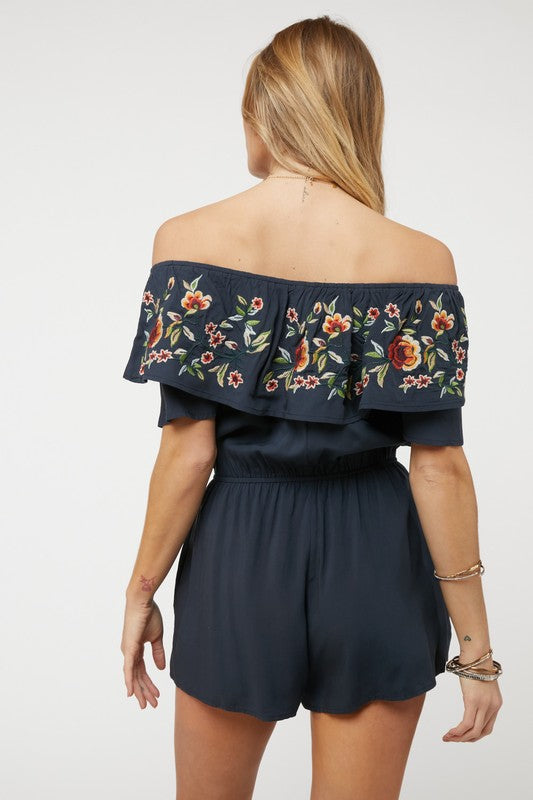 Wearing the stylish Floral Embroidered Off Shoulder Romper, a woman poses gracefully, showcasing its ruffle detail and making it an ideal addition to any summer wardrobe.