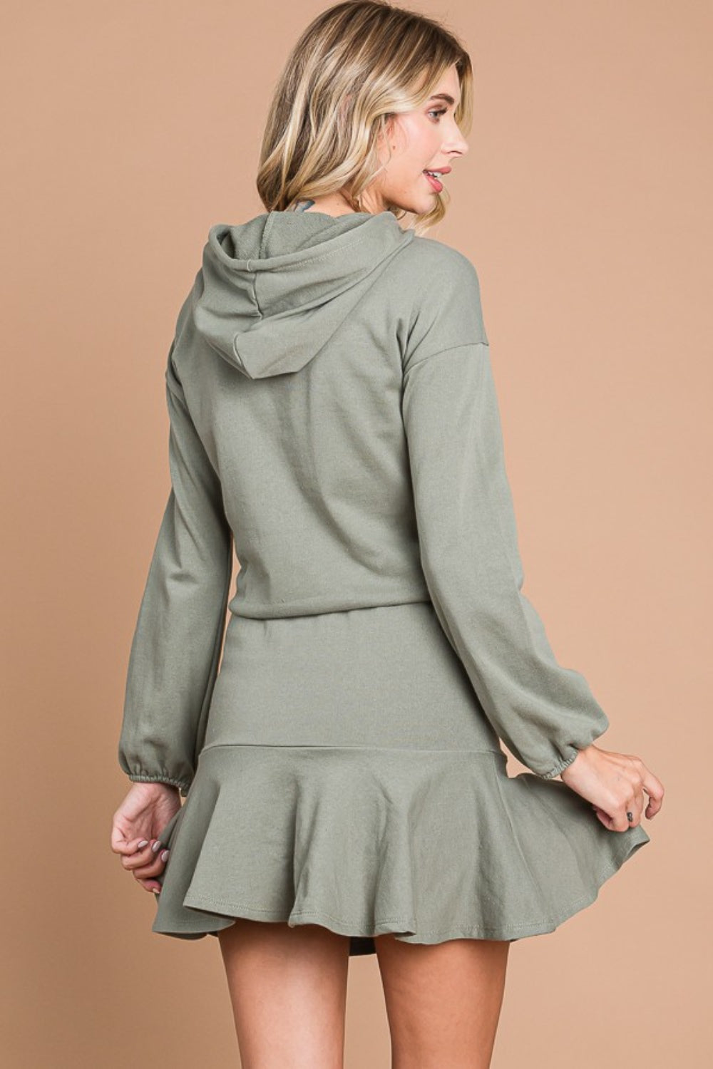 A woman stands smiling against a beige background, wearing the Culture Code Full Size Drawstring Dropped Shoulder Hooded Mini Dress.