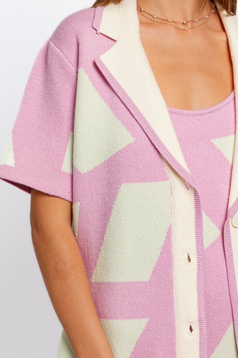 A woman is wearing the Le Lis Abstract Contrast Short Sleeve Collared Cardigan in pastel pink and cream with a geometric pattern, paired with a matching dress. She embodies a modern artistic touch as she looks to the side with her hands at her sides.