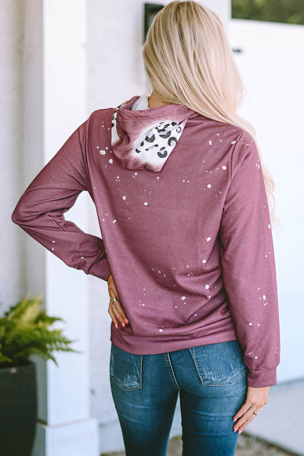 A blonde woman stands with her back to the camera, showcasing a trendy Leopard Tie Dye Print Kangaroo Pocket Pullover Hoodie featuring a maroon design with white speckles. The spotted inner hood lining adds a unique flair, perfectly paired with blue jeans.