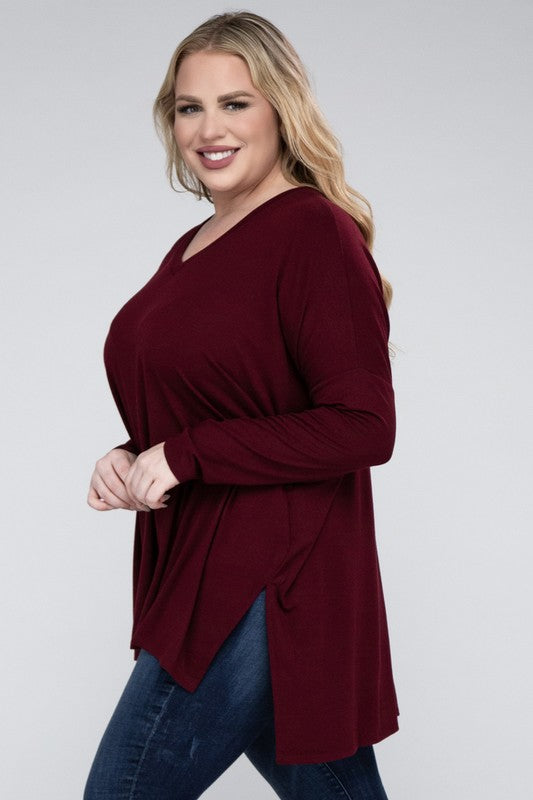 A woman with long, blonde hair wears a Plus Dolman Sleeve V-Neck Side Slit Hi-Low Hem Top and blue jeans, posing with one hand on her hip and smiling at the camera.