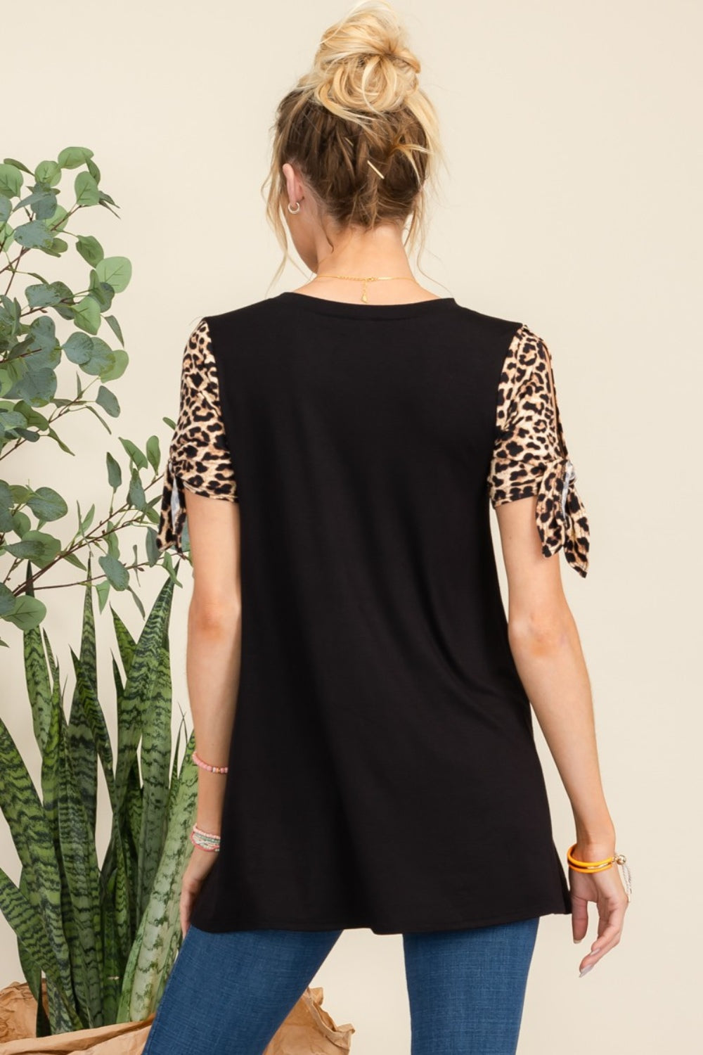 A woman wearing the Celeste Full Size Open Tie Sleeve Leopard Color Blocked Top pairs it effortlessly with blue jeans as she stands near a plant.