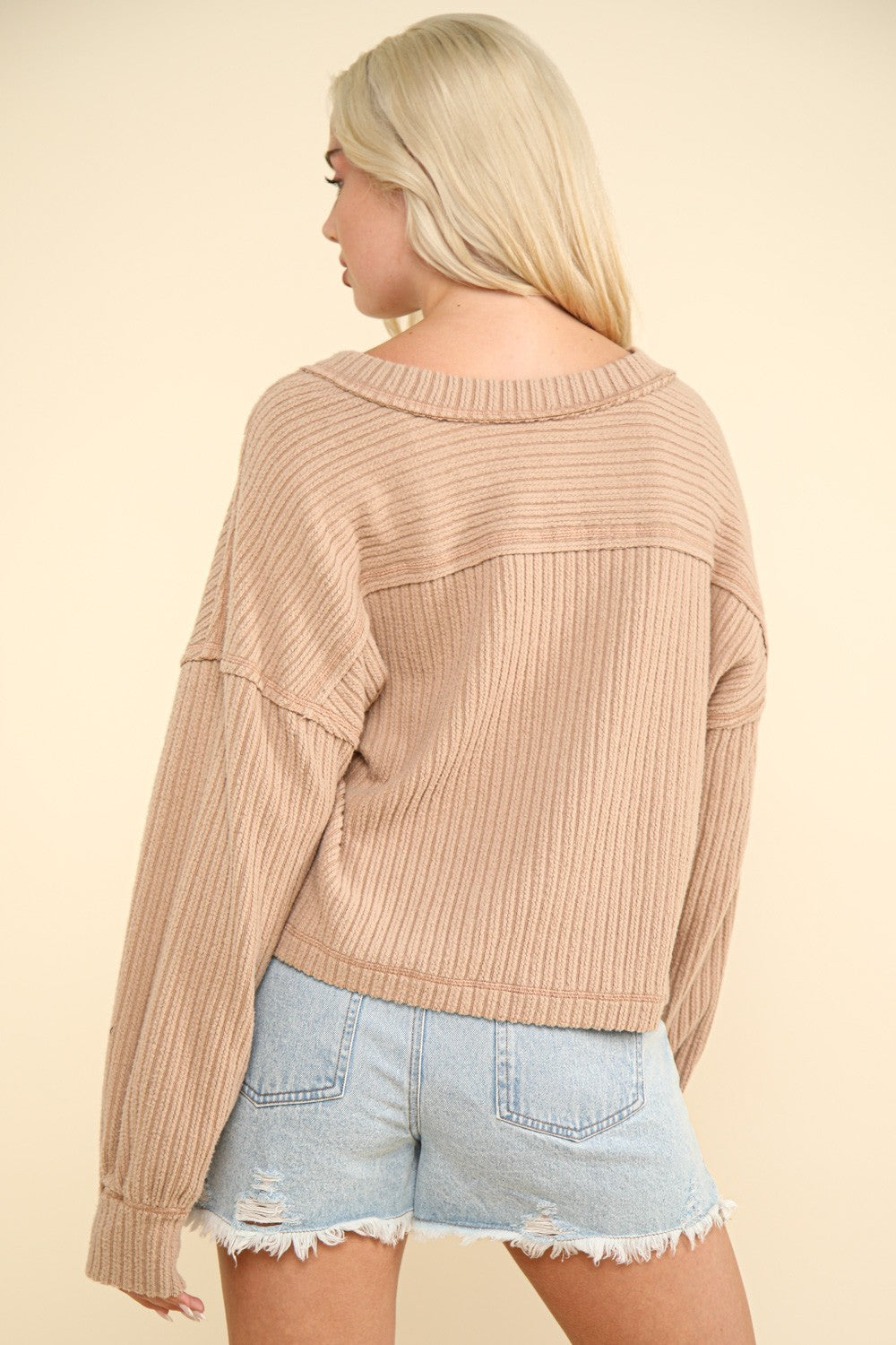 A woman with long blonde hair wears the VERY J Exposed Seam V-Neck Ribbed Knit Top, a casual and comfy piece featuring a ribbed knit texture and a drop shoulder design. She pairs it with light blue ripped denim shorts and stands against a cream-colored background.