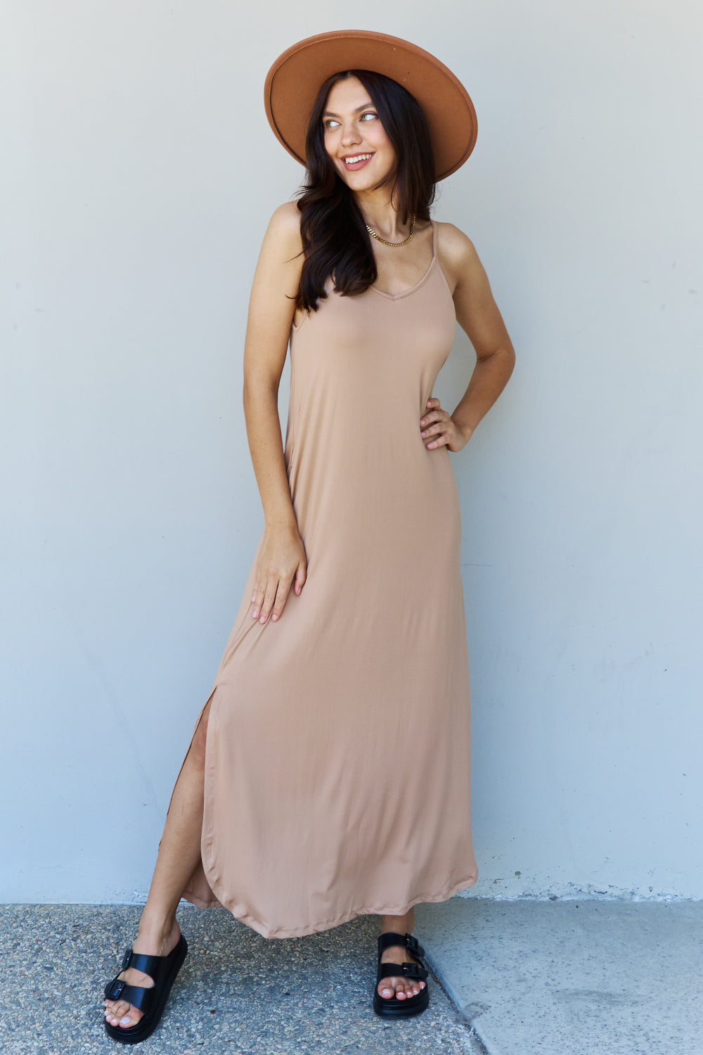 A woman stands against a plain background, wearing the Ninexis Good Energy Full Size Cami Side Slit Maxi Dress in Camel along with a wide-brimmed hat. She has one hand on her hip and is smiling.