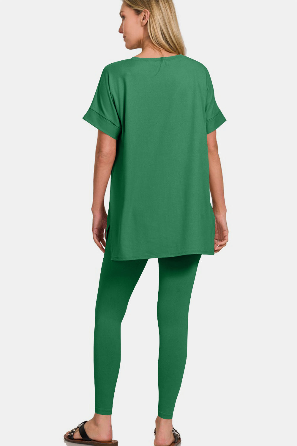 A person is modeling the Zenana Full Size V-Neck Rolled Short Sleeve T-Shirt and Leggings Lounge Set, radiating comfort and style, as they smile and stand with one hand touching their hair.