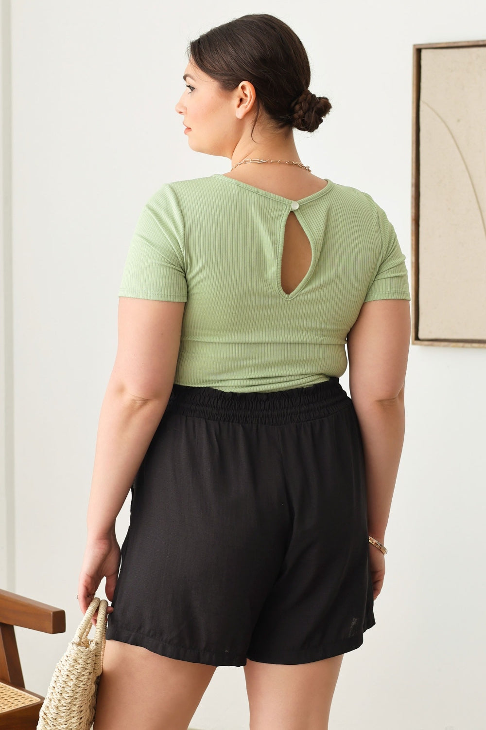 A woman wearing a light green cut-out top and Zenobia Plus Size Drawstring Elastic Waist Shorts with Pockets stands indoors, holding a woven handbag.