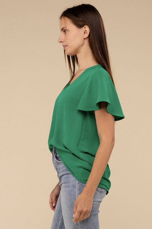 A woman with long straight hair models the Woven Airflow Flutter Sleeve Top in green, matched with light blue jeans, as she poses with her hands in her pockets against a plain beige background.