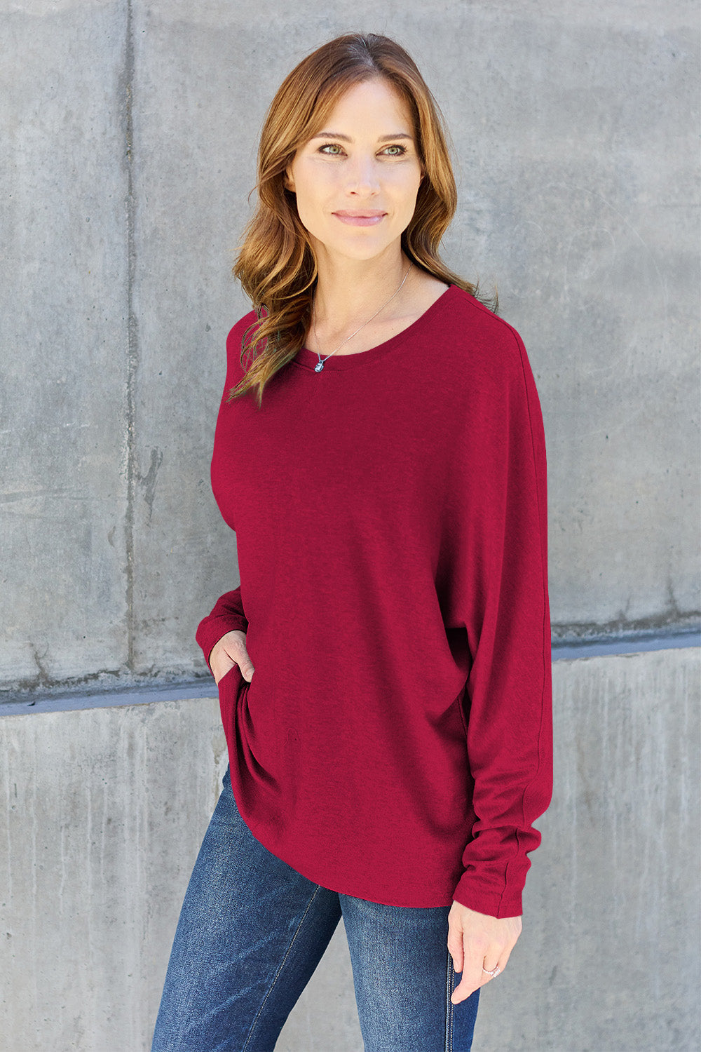 A person wearing a Double Take Full Size Round Neck Long Sleeve T-Shirt in green, blue distressed jeans with a slightly stretchy fit, and a crossbody bag, holding a beverage in a plastic cup.