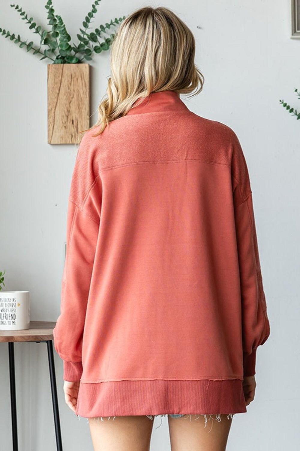 A woman with blonde hair is wearing a First Love Grommet Split Neck Kangaroo Pocket Sweatshirt in coral, standing indoors near a brick wall and plants.