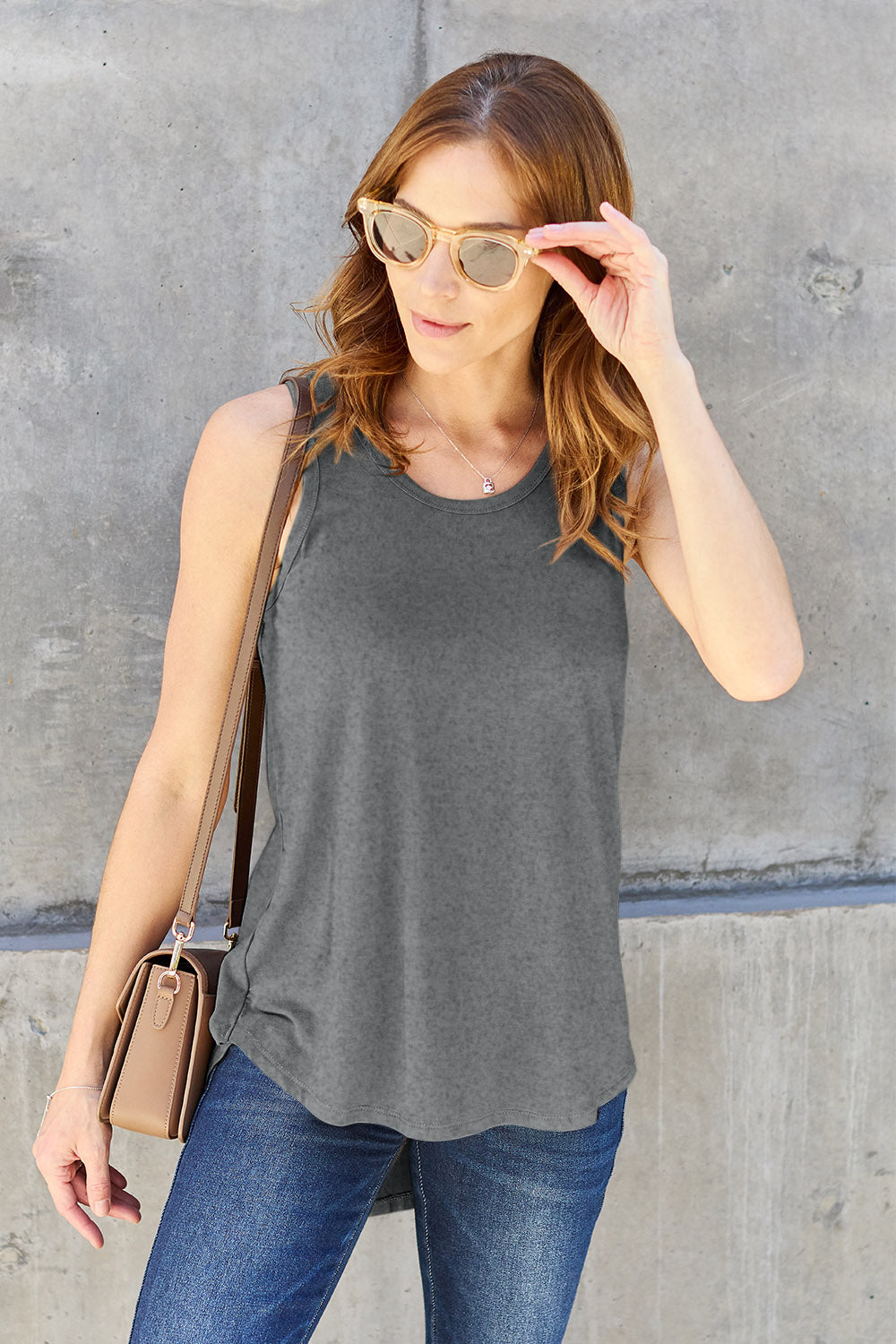 A woman wearing sunglasses, blue jeans, and the Basic Bae Full Size Round Neck Curved Hem Tank is standing in front of a concrete wall, holding her glasses with one hand and carrying a beige shoulder bag. Her basic style complements the slightly stretchy fabric of her outfit, ensuring comfort while maintaining chic simplicity.