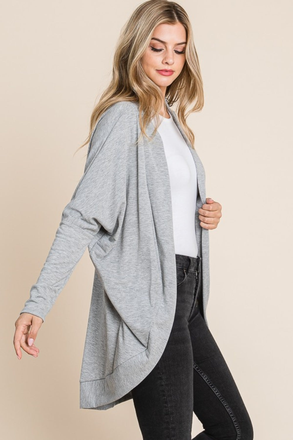 A woman wearing a white shirt, black jeans, and a versatile layering piece—the BOMBOM Open Front Long Sleeve Cocoon Cardigan in grey—stands against a plain beige background.