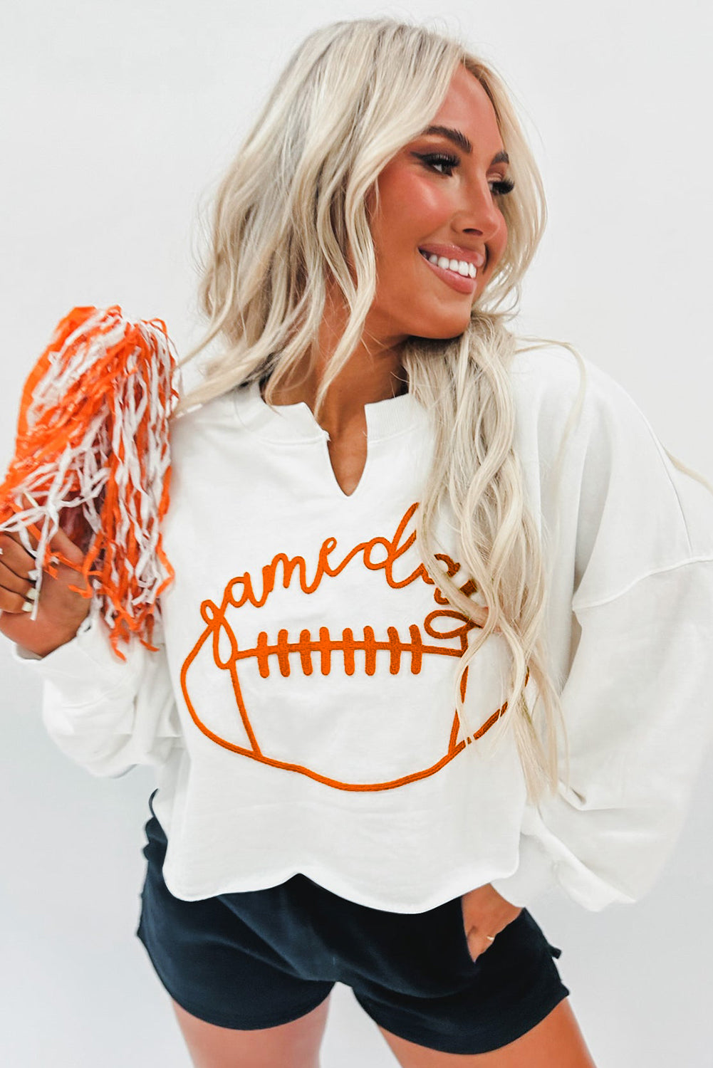 A woman with long blonde hair dressed in a chic White Game Day Lettering Rugby Football Notched Neck Sweatshirt and blue jeans stands near a Christmas tree adorned with red and white ornaments and ribbon.