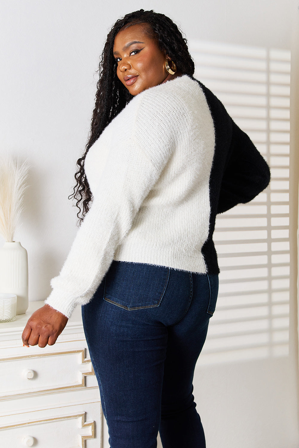 A woman stands with one hand on her hip and the other touching her face. She is wearing the Angel Wings Woven Right Contrast Button-Front V-Neck Cardigan in black and white, paired with jeans. She is smiling.