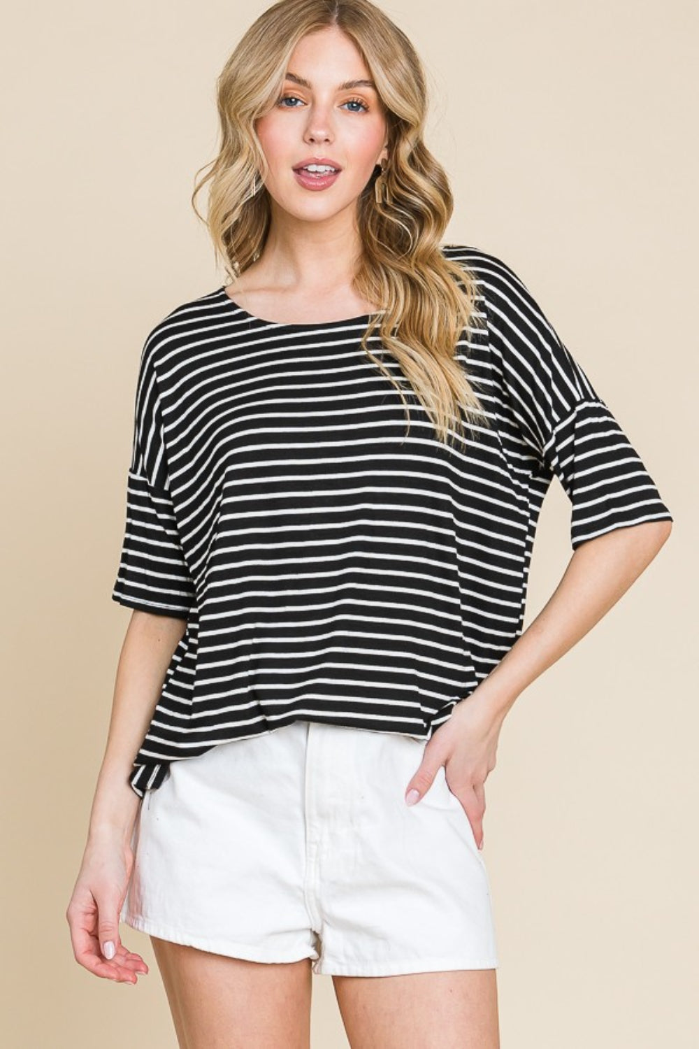 A woman with wavy blonde hair wears a BOMBOM Striped Round Neck T-Shirt and white shorts, standing against a beige background. This classic and casual look highlights her effortless style.