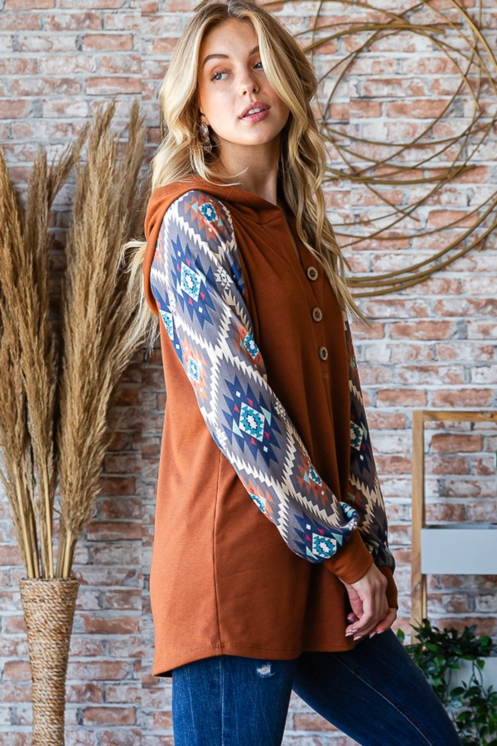 A woman in a Heimish Full Size Half Button Printed Long Sleeve Hooded Top poses casually in front of a brick wall, holding the hood with one hand. She is wearing blue jeans and standing near dried plants, showcasing the unique print design that adds an on-trend touch to her outfit.