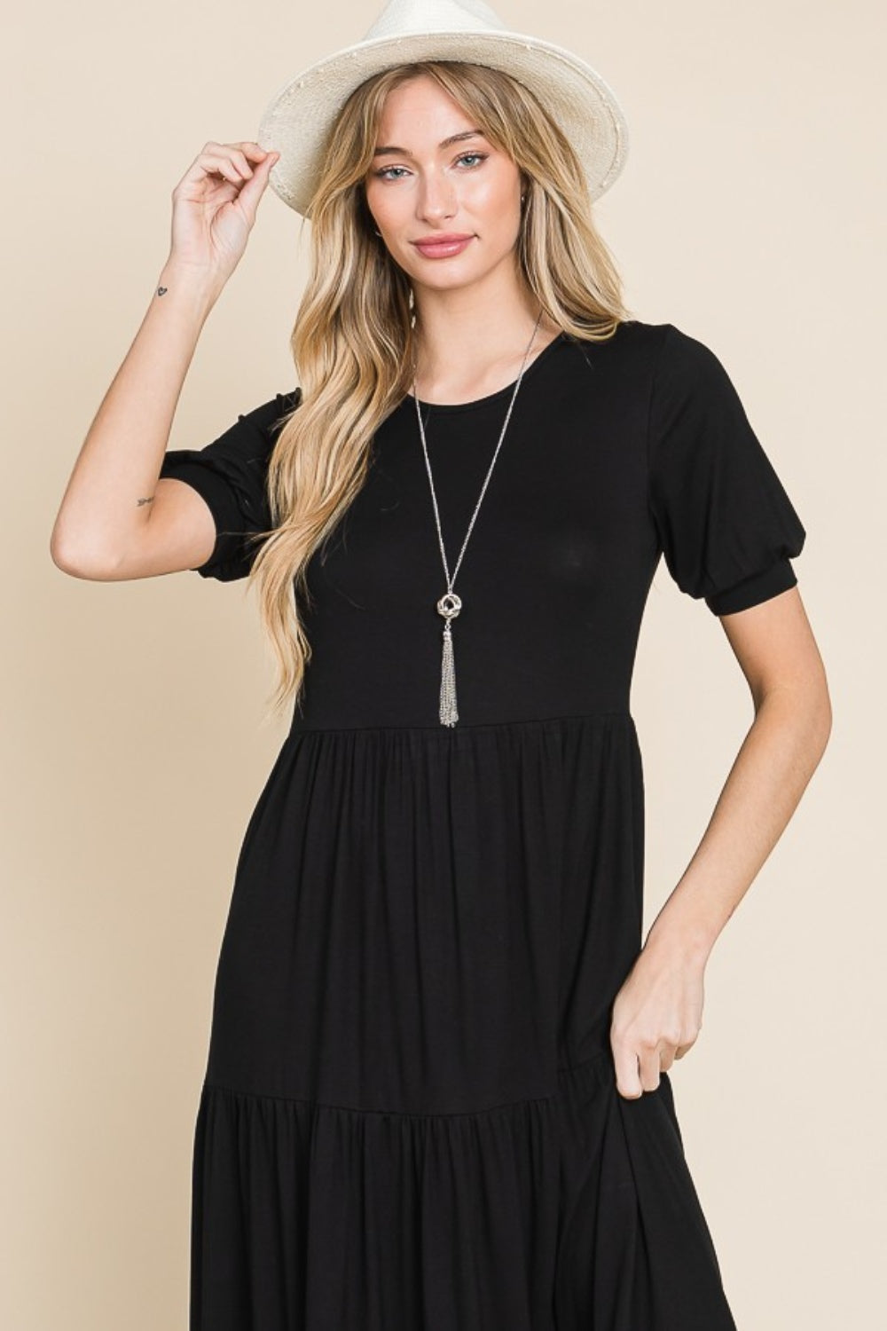 A person wearing the BOMBOM Short Sleeve Tiered Maxi Dress in sleek black stands against a plain beige background, exuding comfort with style. With one hand on their hip and donning white sandals, they effortlessly showcase this versatile piece.