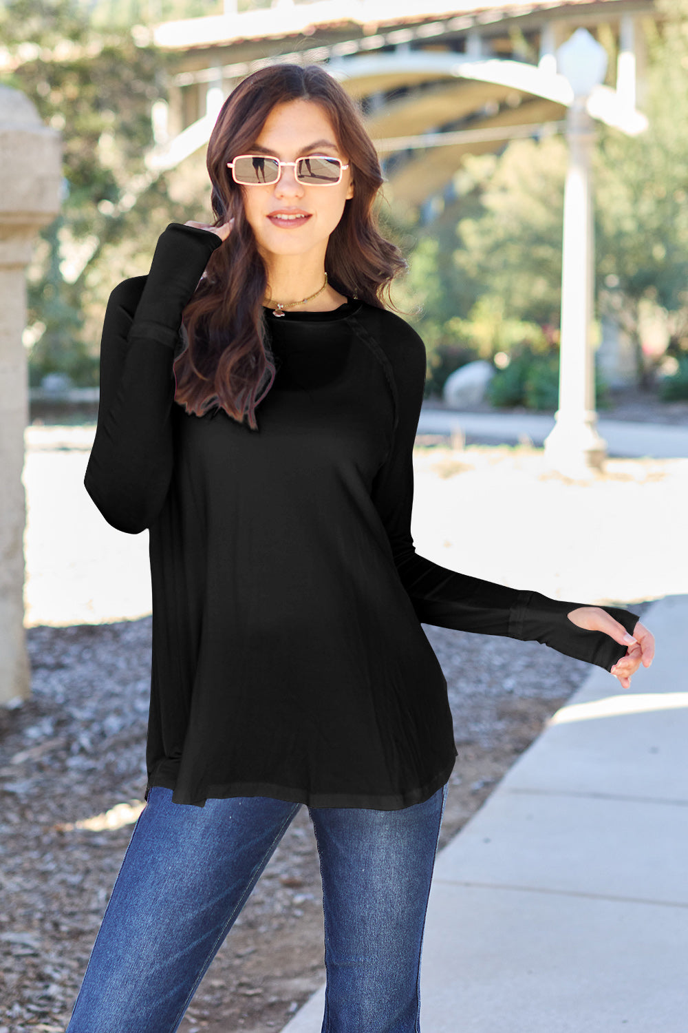 A woman wearing sunglasses and a slightly stretchy Basic Bae Full Size Round Neck Long Sleeve T-Shirt in white stands on a sidewalk with an arched structure in the background, smiling slightly.