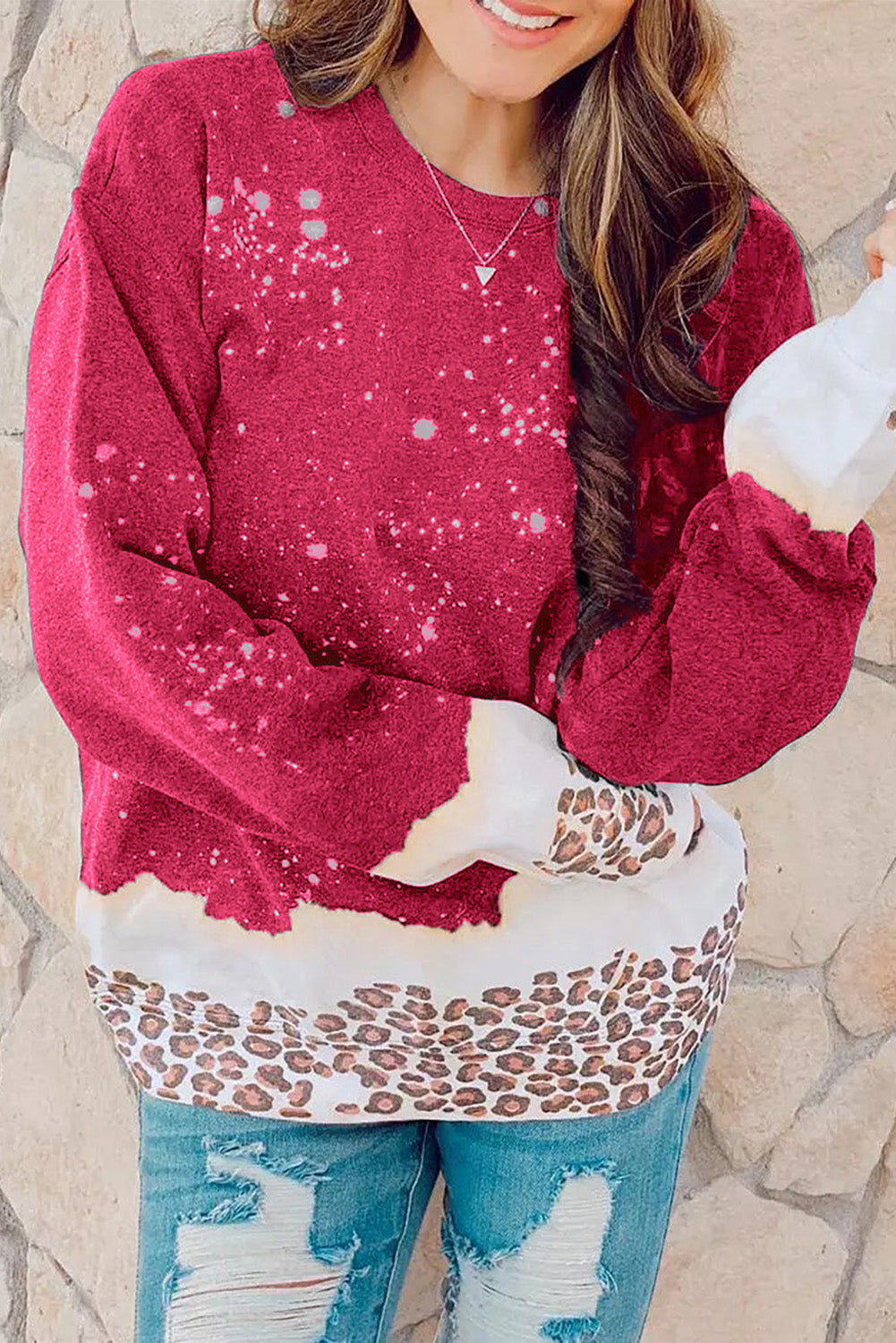 A person with wavy hair is wearing a Fiery Red Leopard Bleached Pullover Sweatshirt paired with blue jeans, standing against a gray background.