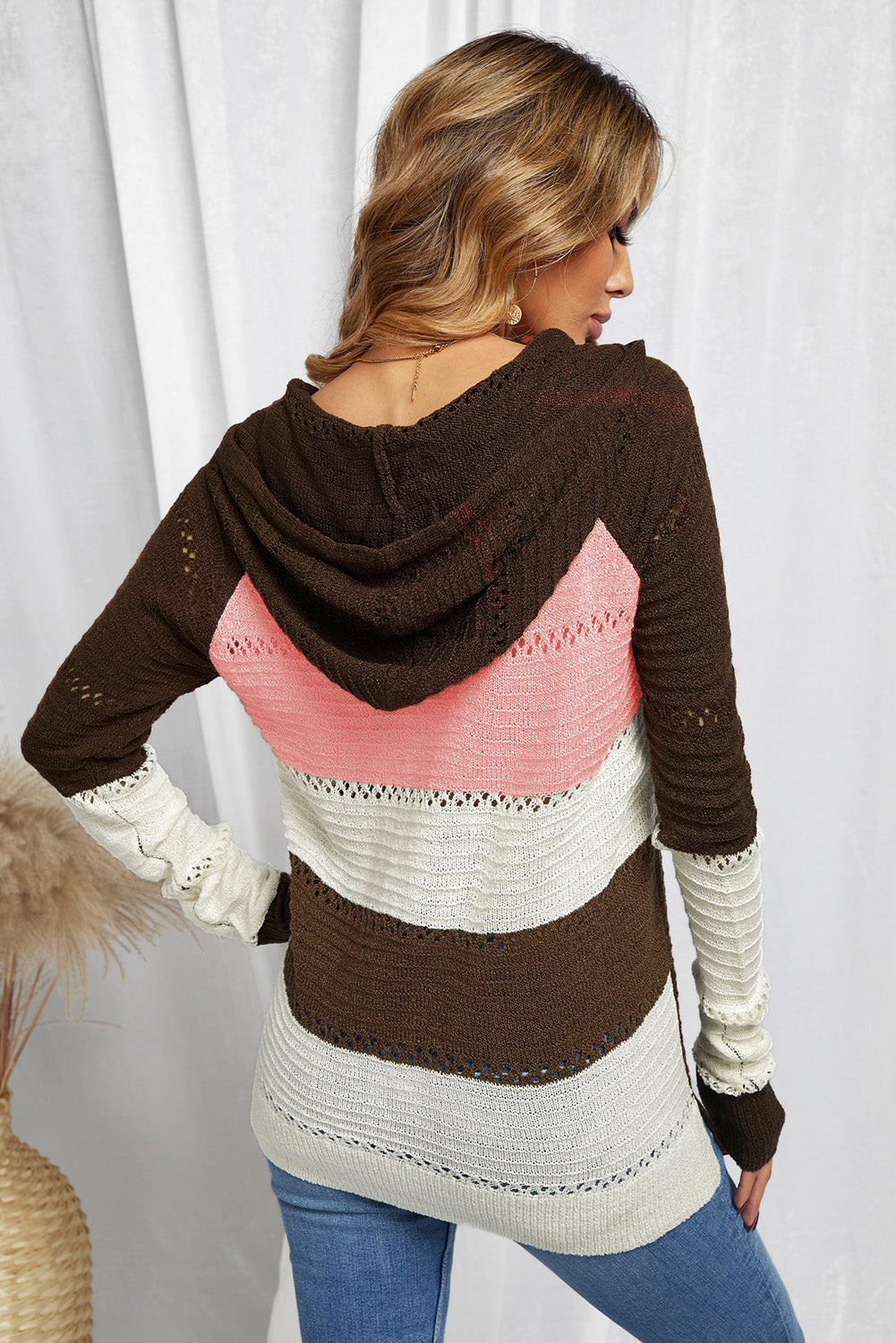 A person stands with their back to the camera, adjusting the hood of a Brown Beach Bonfire Knitted Hoodie featuring dark brown, pink, white, and light brown stripes. Perfect for a casual style, they are wearing distressed blue jeans and showcasing the laid-back vibe of lightweight hoodies.