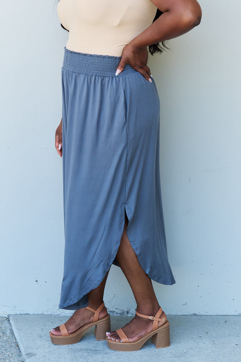 A person wearing a white sleeveless top and the Doublju Comfort Princess Full Size High Waist Scoop Hem Maxi Skirt in Charcoal stands against a plain wall. They are also wearing black sandals.