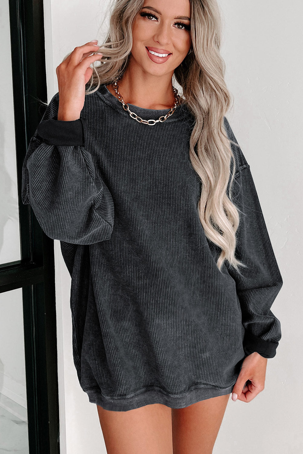 A woman with long blonde hair is smiling and wearing an oversized Black Solid Ribbed Knit Round Neck Pullover Sweatshirt and a gold chain necklace. Showcasing a versatile style, she is standing indoors near a window.