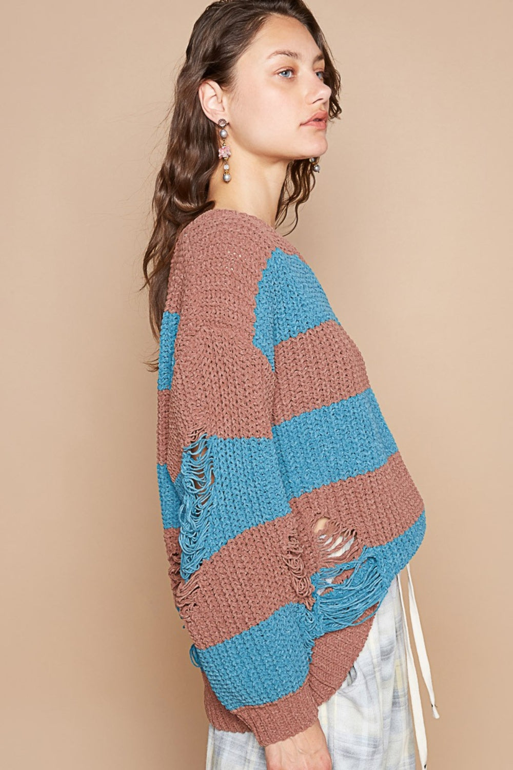 A person with wavy hair is wearing a POL Striped Distressed Long Sleeve Sweater in brown and blue chenille material, paired with light-colored pants, standing against a plain beige background.